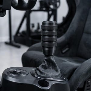 May include: A black cylindrical gear shifter knob with a textured, grooved surface attached to a black leather-like boot. The shifter is mounted on a black plastic base featuring a circular button with a logo and a small circular indentation.