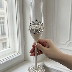 Puede incluir: Una copa de champ&aacute;n de cristal transparente con tallo y base de color blanco texturizado. La base est&aacute; decorada con una disposici&oacute;n circular de perlas blancas y cristales transparentes. Una banda decorativa a juego rodea la parte inferior.