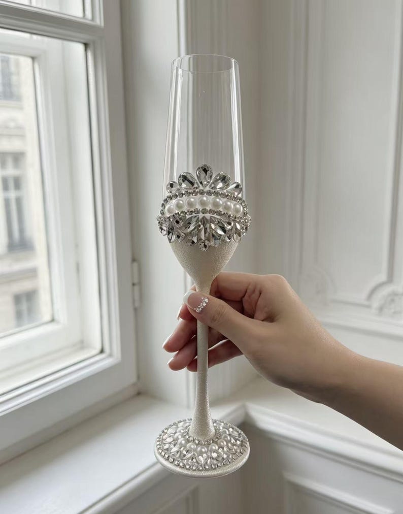 Puede incluir: Una copa de champ&aacute;n de cristal transparente con tallo y base de color blanco texturizado. La base est&aacute; decorada con una disposici&oacute;n circular de perlas blancas y cristales transparentes. Una banda decorativa a juego rodea la parte inferior.