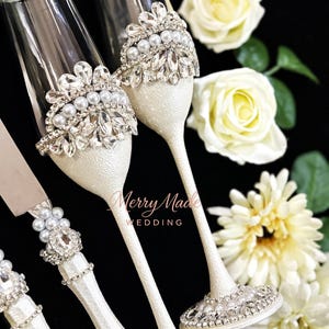 Puede incluir: Dos copas de champ&aacute;n de cristal transparente y un cuchillo para pasteles con tallos blancos texturizados. Cada pieza est&aacute; decorada con filas de perlas blancas y cristales transparentes facetados en forma de corona. El texto Merry Made WEDDING es visible.
