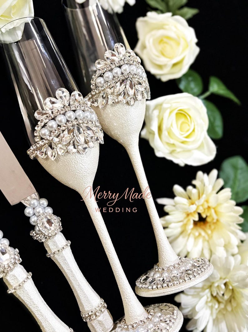 Puede incluir: Dos copas de champ&aacute;n de cristal transparente y un cuchillo para pasteles con tallos blancos texturizados. Cada pieza est&aacute; decorada con filas de perlas blancas y cristales transparentes facetados en forma de corona. El texto Merry Made WEDDING es visible.