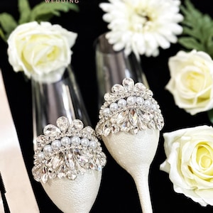 Puede incluir: Dos copas de champ&aacute;n con tallos y bases de textura blanca, adornadas con coronas plateadas con cristales facetados transparentes y perlas blancas. El texto Merry Made WEDDING es visible en la esquina superior izquierda de la imagen.