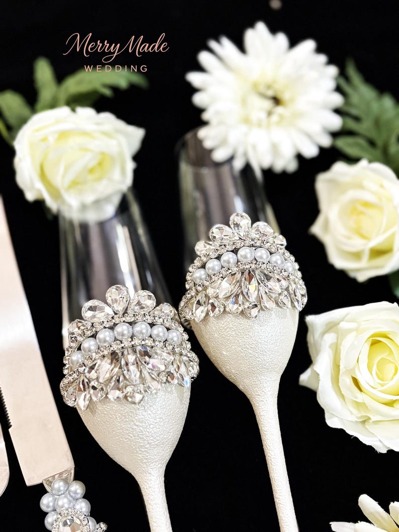 Puede incluir: Dos copas de champ&aacute;n con tallos y bases de textura blanca, adornadas con coronas plateadas con cristales facetados transparentes y perlas blancas. El texto Merry Made WEDDING es visible en la esquina superior izquierda de la imagen.