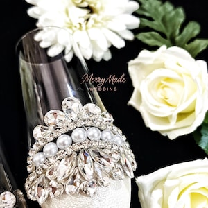 Puede incluir: Una copa de champ&aacute;n de cristal transparente decorada con un adorno plateado en forma de corona con cristales facetados transparentes y una fila de perlas blancas. El texto Merry Made WEDDING est&aacute; impreso en cursiva sobre el cristal.