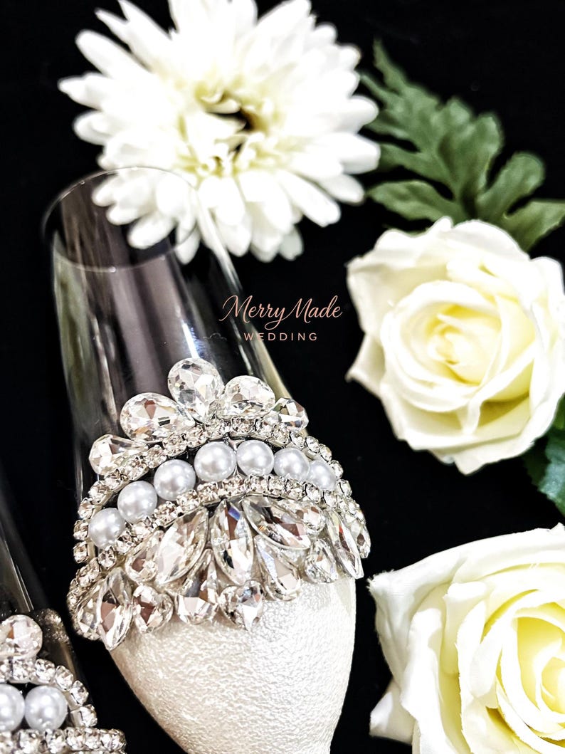 Puede incluir: Una copa de champ&aacute;n de cristal transparente decorada con un adorno plateado en forma de corona con cristales facetados transparentes y una fila de perlas blancas. El texto Merry Made WEDDING est&aacute; impreso en cursiva sobre el cristal.