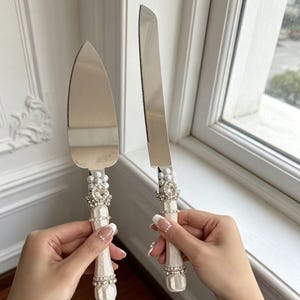 Puede incluir: Un servidor de pasteles y un cuchillo para pasteles a juego en color plata con mangos blancos texturizados. Cada mango est&aacute; decorado con un grupo de perlas blancas falsas, un brillante central transparente y bandas de cristales peque&ntilde;os.