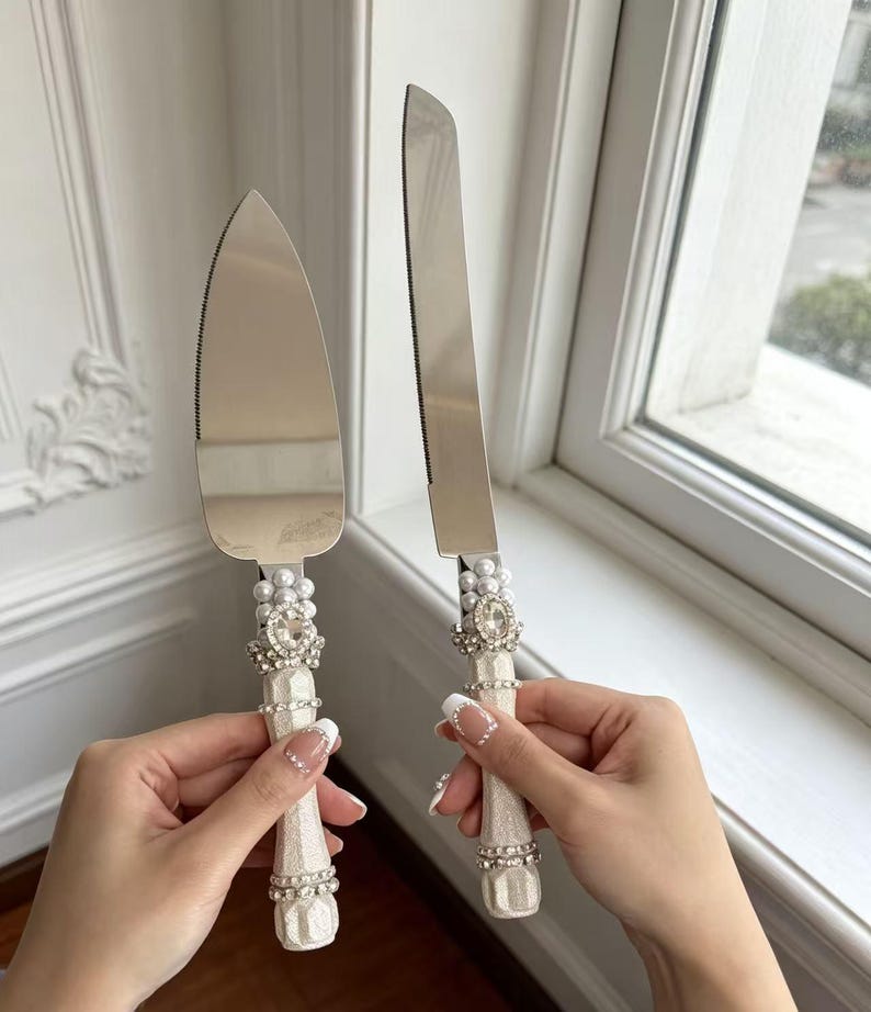 Puede incluir: Un servidor de pasteles y un cuchillo para pasteles a juego en color plata con mangos blancos texturizados. Cada mango est&aacute; decorado con un grupo de perlas blancas falsas, un brillante central transparente y bandas de cristales peque&ntilde;os.