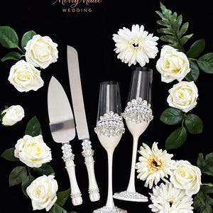 Puede incluir: Un juego de cuchillo y pala para pastel junto a dos copas de champ&aacute;n, con mangos y tallos blancos decorados con pedrer&iacute;a transparente y perlas falsas. El texto Merry Made WEDDING aparece arriba. Rosas y margaritas blancas rodean los objetos.