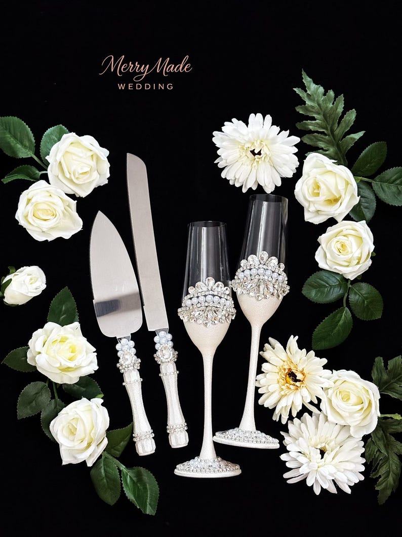 Puede incluir: Un juego de cuchillo y pala para pastel junto a dos copas de champ&aacute;n, con mangos y tallos blancos decorados con pedrer&iacute;a transparente y perlas falsas. El texto Merry Made WEDDING aparece arriba. Rosas y margaritas blancas rodean los objetos.