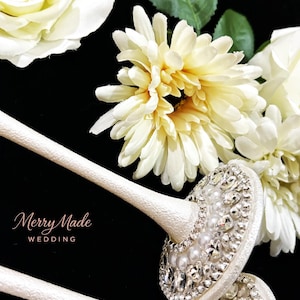 Puede incluir: Dos tallos texturizados de color blanco roto con partes superiores circulares adornadas con pedrer&iacute;a transparente y perlas blancas. El texto Merry Made WEDDING est&aacute; impreso en dorado. Rosas y margaritas artificiales color crema al fondo.