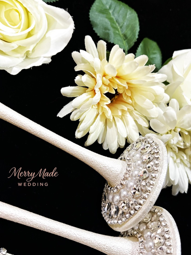 Puede incluir: Dos tallos texturizados de color blanco roto con partes superiores circulares adornadas con pedrer&iacute;a transparente y perlas blancas. El texto Merry Made WEDDING est&aacute; impreso en dorado. Rosas y margaritas artificiales color crema al fondo.