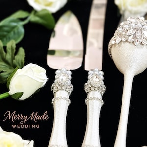 Puede incluir: Un juego de cuchillo y pala para pastel en plata con mangos blancos texturizados, decorados con filas de pedrer&iacute;a transparente, perlas artificiales y un cristal ovalado central. Se incluye una copa blanca a juego con cristales y perlas.