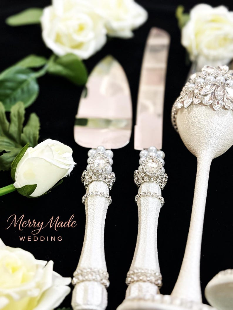 Puede incluir: Un juego de cuchillo y pala para pastel en plata con mangos blancos texturizados, decorados con filas de pedrer&iacute;a transparente, perlas artificiales y un cristal ovalado central. Se incluye una copa blanca a juego con cristales y perlas.