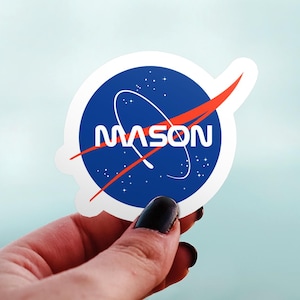 May include: A white sticker with a blue circle design, a red swoosh, and the name "MASON" in white letters. The sticker is held up against a light blue background.