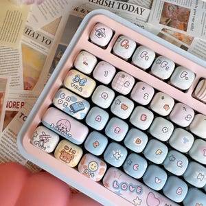 May include: A pink and blue keyboard with custom keycaps featuring cartoon characters and designs. The keycaps have words like "Tab", "Shift", "Win", "Alt", "Love", and "Happy" printed on them.