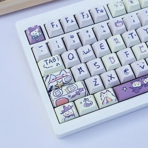 May include: A white mechanical keyboard with custom keycaps featuring cartoon cat illustrations in pastel colors. The keycaps have various designs, including cats in witch hats, bats, and pumpkins. The keyboard has standard letter and number keys.