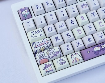 May include: A white mechanical keyboard with custom keycaps featuring cartoon cat illustrations in pastel colors. The keycaps have various designs, including cats in witch hats, bats, and pumpkins. The keyboard has standard letter and number keys.
