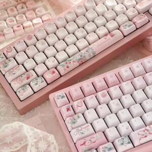 May include: Two pink mechanical keyboards with floral and pastel-colored keycaps. The keyboards have a white base and feature a floral design on the space bar.