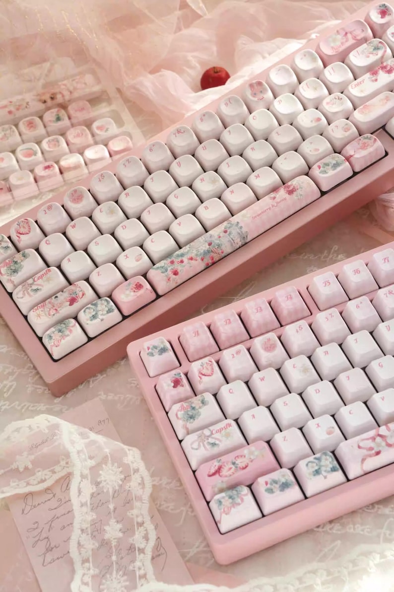 May include: Two pink mechanical keyboards with floral and pastel-colored keycaps. The keyboards have a white base and feature a floral design on the space bar.