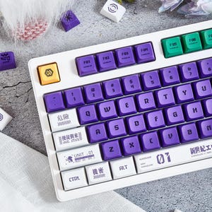 May include: A white keyboard with purple keys and white keycaps. The keycaps have Japanese and English text, including "Angel Attack" and "Unit 01".