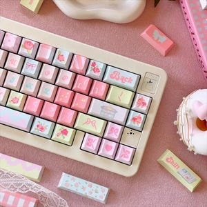 May include: A white and pink keyboard with pastel-coloured keycaps featuring floral and bow designs. The keyboard has a 'Back' key and a 'Dino' key. The keyboard is surrounded by pastel-coloured accessories, including a donut and a small pink and white box.