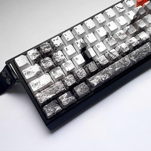 Puede incluir: Un teclado mecánico negro con teclas personalizadas. Las teclas alternan entre blanco y negro, con un diseño veteado o texturizado único. Algunas teclas son blancas con letras negras, otras son negras con letras blancas. El teclado tiene un reposamuñecas de cuero.