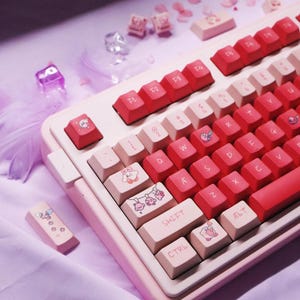 May include: A custom mechanical keyboard in pink and white. The keycaps are red and white, featuring cartoon character designs. The keyboard has a standard layout with function keys and a shift key. The keyboard is on a purple surface.