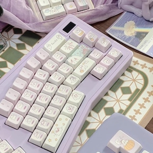 May include: A lilac mechanical keyboard with white keycaps decorated with cartoon ghost and pumpkin illustrations. The keyboard is rectangular with a number pad and a black power button. The keycaps are pale pink and arranged in a standard QWERTY layout.