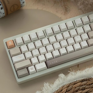 May include: A white and beige mechanical keyboard with the word "DUNE" printed on the space bar. The keyboard has a light green frame and white keys with black lettering. The keys are arranged in a standard QWERTY layout.