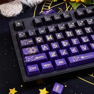 May include: A purple and black mechanical keyboard with gold keycaps featuring a celestial theme. The keyboard has a black and white starry background with a crescent moon and stars. The keycaps have runes and symbols on them.