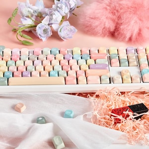 May include: A white keyboard with pastel colored keycaps. The keyboard has a white frame and a white space bar. The keycaps are in various shades of pink, blue, yellow, and green. The keyboard is sitting on a white surface.