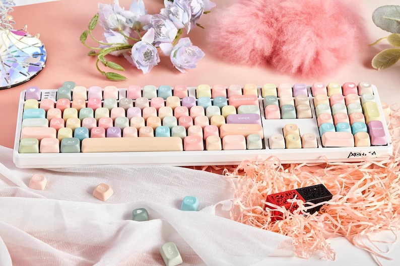 May include: A white keyboard with pastel colored keycaps. The keyboard has a white frame and a white space bar. The keycaps are in various shades of pink, blue, yellow, and green. The keyboard is sitting on a white surface.