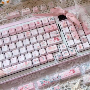 May include: A pink and white keyboard with custom keycaps featuring floral and cartoon designs. The keyboard has a pink frame and white keys. The keycaps have a variety of designs, including flowers, strawberries, and cartoon characters.