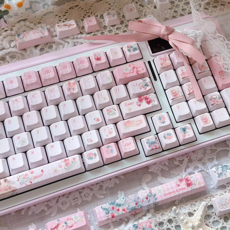 May include: A pink and white keyboard with custom keycaps featuring floral and cartoon designs. The keyboard has a pink frame and white keys. The keycaps have a variety of designs, including flowers, strawberries, and cartoon characters.