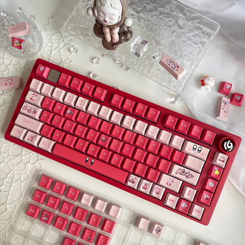 May include: A red mechanical keyboard with white and pink keycaps. The keyboard has custom keycaps with cartoon character designs. Additional keycaps are visible in a clear plastic case. A small figurine and decorative elements are also present.