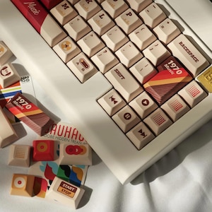 Può includere: Una tastiera meccanica bianca con tasti marroni e rossi. I tasti hanno diversi design, tra cui le parole "Music", "1970" e "IBM".