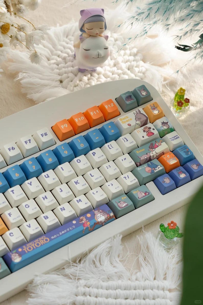 May include: A white mechanical keyboard with blue and orange keys. The keys have various illustrations of characters from the animated film My Neighbor Totoro. The keyboard has a blue and white space bar with the text "Mio Vicino Totoro" and Japanese characters.