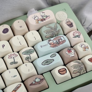 Può includere: Una tastiera verde e crema con tasti personalizzati che presentano illustrazioni di fiori, foglie e animali. I tasti sono realizzati in plastica bianca liscia e hanno una finitura opaca. La tastiera ha un layout standard con una riga di numeri, una riga di funzioni e un'area di tastiera principale. I tasti sono disposti in uno schema sfalsato, con i tasti più grandi nella riga inferiore e i tasti più piccoli nella riga superiore. La tastiera è collegata a un computer tramite un cavo USB.