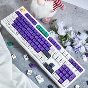 May include: A white mechanical keyboard with purple and green keycaps. The keyboard has a Japanese inscription that reads "新世紀エヴァンゲリオン" which translates to "Neon Genesis Evangelion".