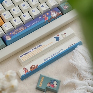 May include: A white keyboard with blue keys and a blue and white keyboard keycap set featuring a character from the movie My Neighbor Totoro. The keycap set includes the text "My Neighbor Totoro" and "Il Mio Vicino Totoro" in Japanese. The keycap set also includes the text "温めらゑがざむり" in Japanese. The keyboard is sitting on a white fluffy surface.