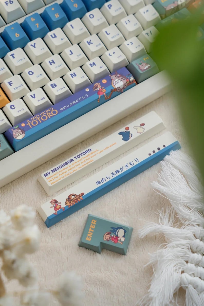 May include: A white keyboard with blue keys and a blue and white keyboard keycap set featuring a character from the movie My Neighbor Totoro. The keycap set includes the text "My Neighbor Totoro" and "Il Mio Vicino Totoro" in Japanese. The keycap set also includes the text "温めらゑがざむり" in Japanese. The keyboard is sitting on a white fluffy surface.