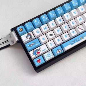 May include: A black mechanical keyboard with custom keycaps featuring a blue and white color scheme and illustrations of the character Doraemon. The keyboard has a wrist rest and a variety of keycap designs.