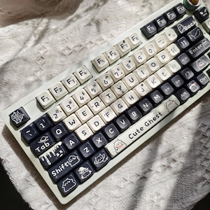 May include: A custom mechanical keyboard with white and black keycaps. The keycaps feature various icons and the words "Cute Ghost". The keyboard has a light green frame and is resting on a white, textured surface.