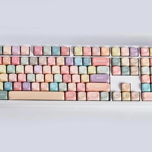 May include: A pastel-colored keyboard with white lettering on the keys. The keys are arranged in a standard QWERTY layout. The keyboard has a white frame and a light pink space bar.