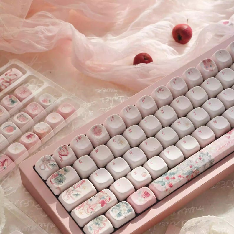 May include: A pink mechanical keyboard with white keycaps and floral designs. The keyboard has a pink frame and is surrounded by pink fabric.