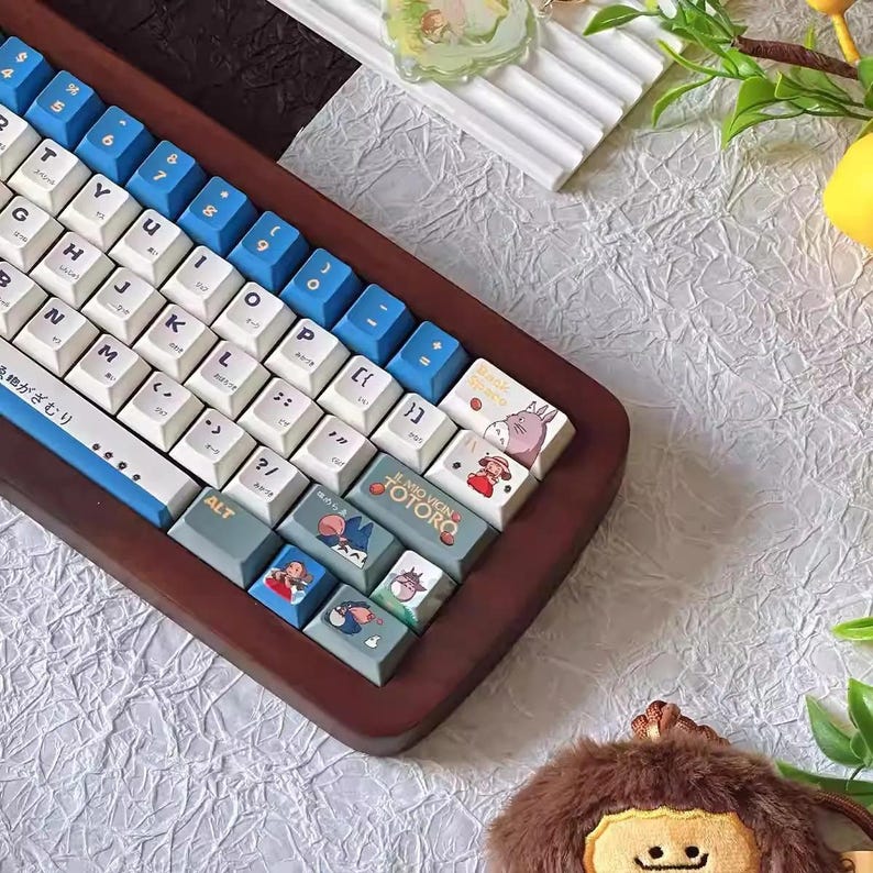 May include: A wooden keyboard with blue and white keys and custom keycaps featuring characters from the animated film "My Neighbor Totoro". The keycaps have illustrations of Totoro, the cat bus, and other characters from the film. The keyboard is surrounded by a brown wooden frame.