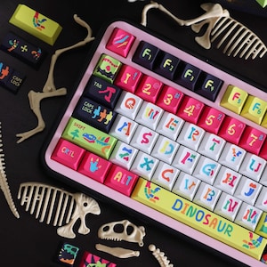 May include: A pink and black keyboard with colourful keycaps featuring dinosaur bones and other dinosaur-themed designs. The keyboard has a space bar with the word "DINOSAUR" printed on it.