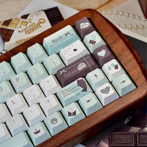 Può includere: Una tastiera blu chiaro e bianca con tasti marroni. I tasti hanno diversi disegni, tra cui una barretta di cioccolato, un cuore e un cupcake. La tastiera è in legno e ha una finitura marrone chiaro. Il testo sui tasti dice "Back X", "Enter" e "Indulge in the crisp coolness of mint chocolate."