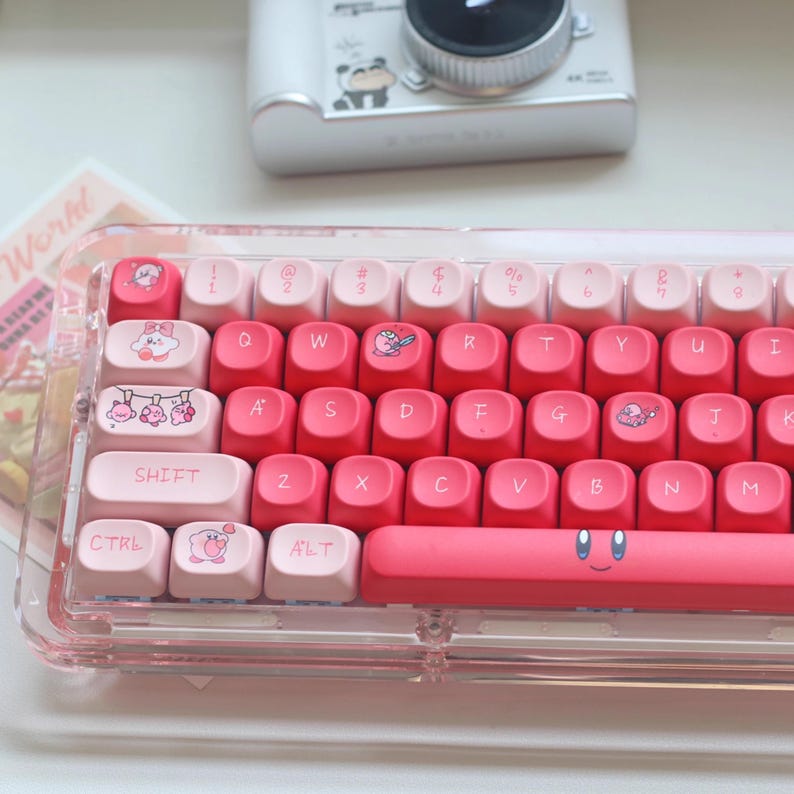 May include: A pink and white mechanical keyboard with custom keycaps. The keycaps feature cartoon character designs and a gradient colour scheme. The keyboard is housed in a clear acrylic case, and the base bar has a smiling face design.