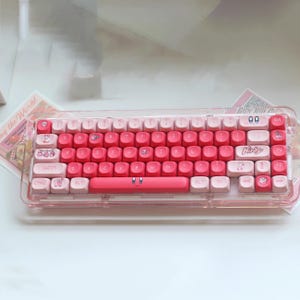 May include: A pink and white mechanical keyboard with custom keycaps. The keyboard has a gradient design, transitioning from white to pink. The keycaps feature character illustrations and the word "Kirby". It is encased in a clear plastic case.